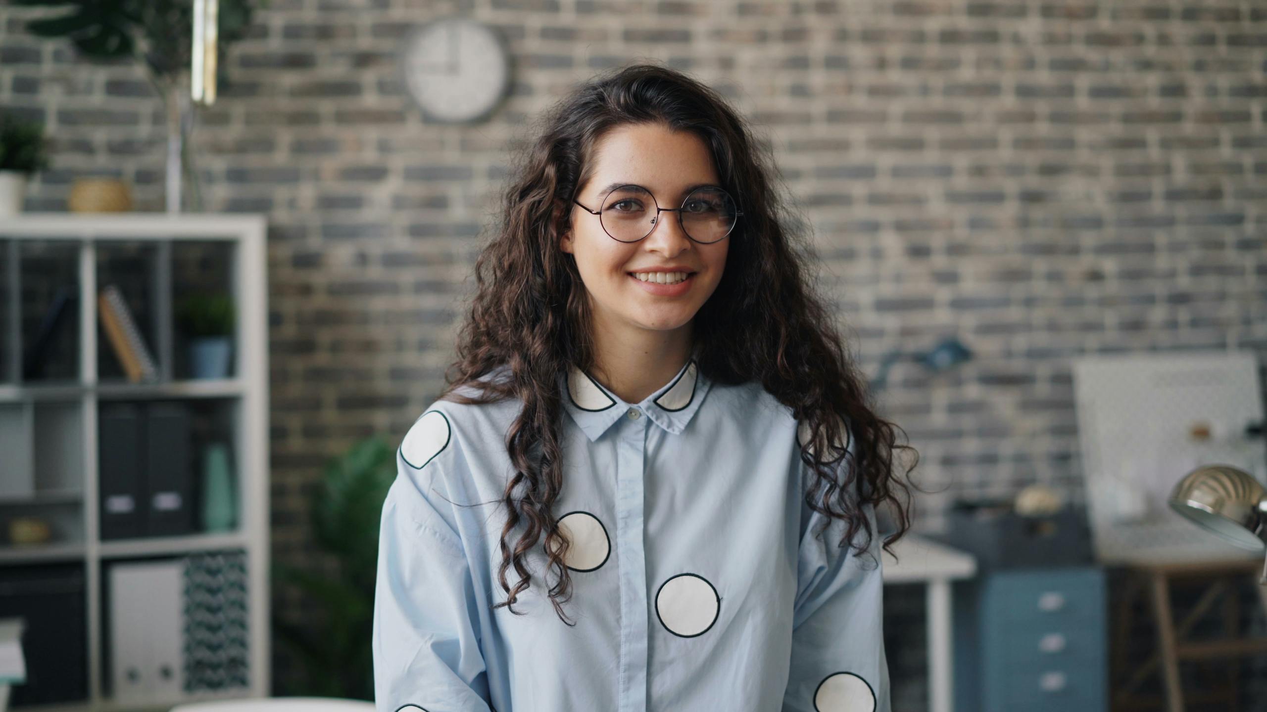 Portrait of a smiling woman with curly hair and eyeglasses in an office setting.