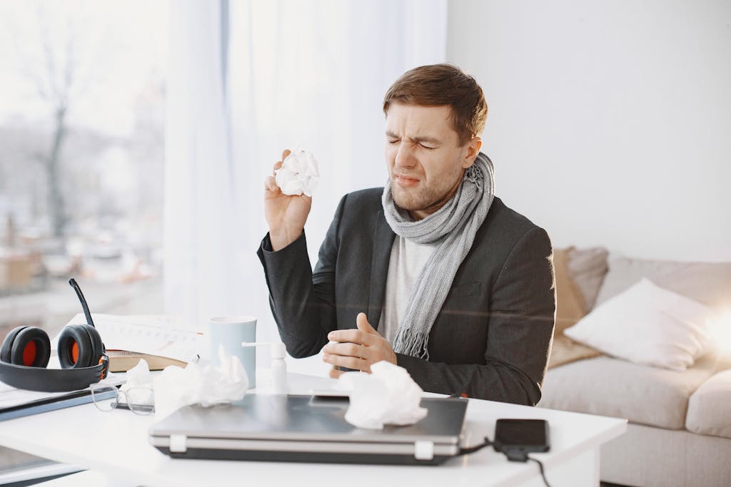 A sick man wearing a scarf sitting at his desk, holding a tissue, displaying signs of discomfort.