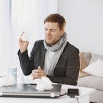 A sick man wearing a scarf sitting at his desk, holding a tissue, displaying signs of discomfort.
