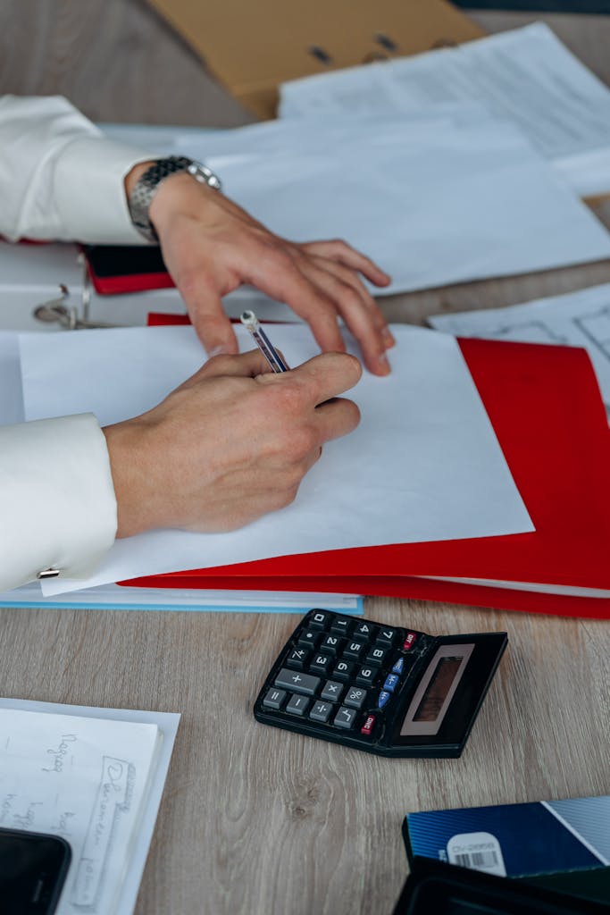Close-up of a businessman writing on paper with a calculator and documents on a wooden desk.