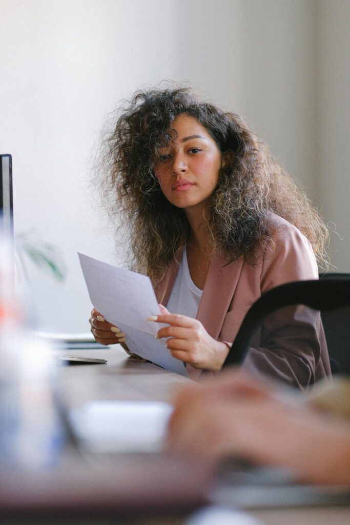 Pensive female with dark hair and paper sheet in hands sitting at table while working in conference room with blurred crop colleague