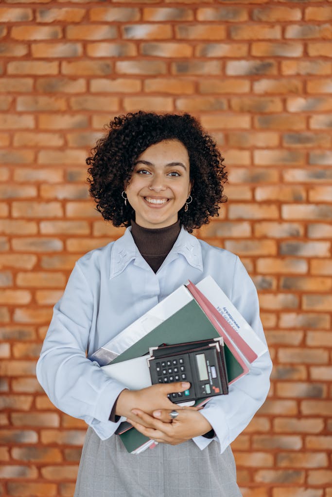Smiling woman with curly hair holding a calculator and folders, standing against a brick wall.